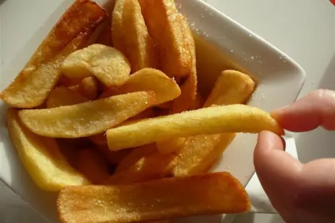 A small portion of golden, fried chips in a square white bowl. One chip is being held between a pink finger and thumb.