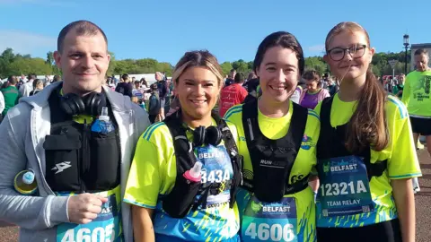 There are four people in the picture. From left to right is a dark haired man with a grey jacket and black running vest. There are three women next to him with a hi-vis top on and black running vest.