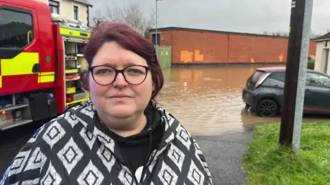 Sarah Kirkwood - a woman with red hair tied up wearing black circle framed glasses and a black and white patterned coat looks at the camera. In the background is a red fire engine parked beside brown flood water. There is a small grey car in the floodwater.