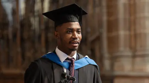 Aaron Agbanu speaks in front of microphone on a lectern. He wears a black graduation hat and robe with a blue caped strap across his tie. He is inside the grand interior of Chester Cathedral.