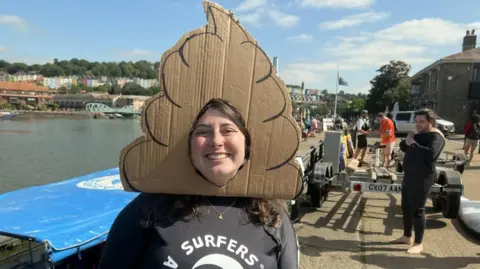 A woman in a black tee-shirt stands at the waters edge of a harbour. Houses and bridges are visible in the background, while other people are walking by the water. The woman is wearing a hat made out of cardboard in the shape of a poo.