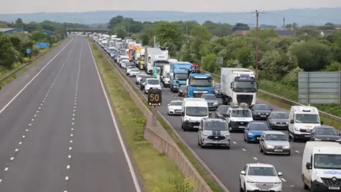 An empty motorway in one direction with large congestion in the other direction, with queues of vehicles all the way along.