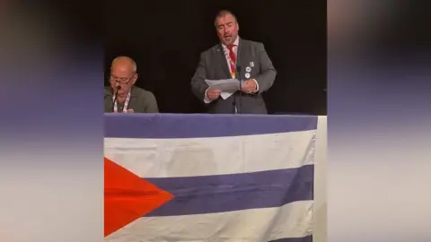Facebook/Embassy of Cuba in the UK Steve Witherden looking down at a speech in front of a Cuban flag.