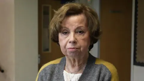 A woman with a brown bob haircut, looking at the camera, wearing a mainly grey cardigan with a yellow stripe down the arms.
