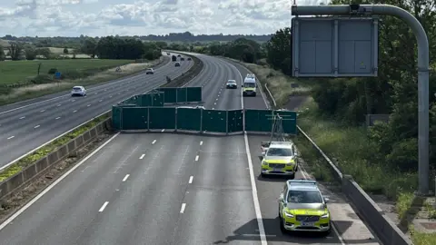 A number of police and highways vehicles are shown parked on the hard shoulder of the motorway, which is completely blocked off by a large temporary screen around two cars. The cars are almost completely hidden from view. 