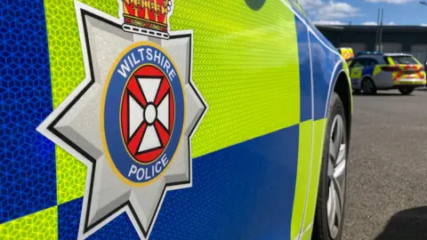 The side of a Wiltshire Police car. It has the Wiltshire police logo on the door. It is a star like shape with a yellow, blue and red circle inside it. It has Wiltshire Police written in white lettering, with a crown on top.
