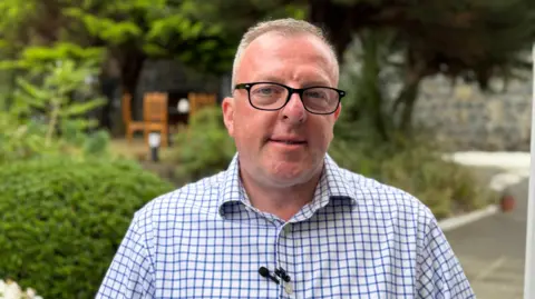 A middle-aged man in a blue and white checked shirt looking at the camera 