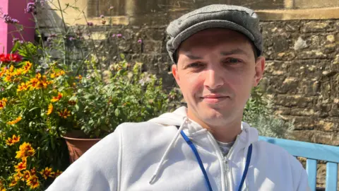 BBC Damian Millard smiling at the camera, wearing a white zip-up hoodie, a blue lanyard, and a grey flat cap. He is sitting on a blue bench outside, surrounded by yellow and orange flowers in plant pots. 