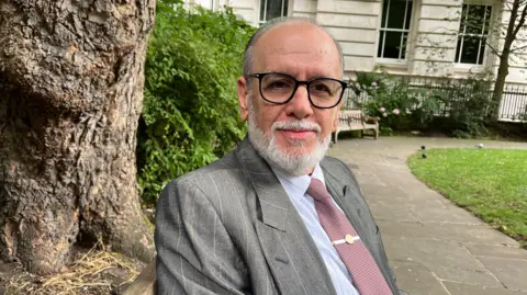 George Psaradakis in a mauve tie, grey suit and black glasses sits on a bench in a park with a tree trunk behind him.