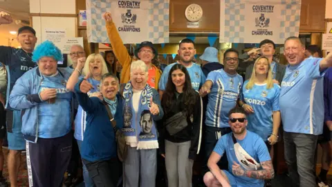 Staff and visitors at the Unicorn Social Club, many wearing Coventry City tops. A man on the left is wearing a curly light blue wig and several people are raising the arms in celebration. 