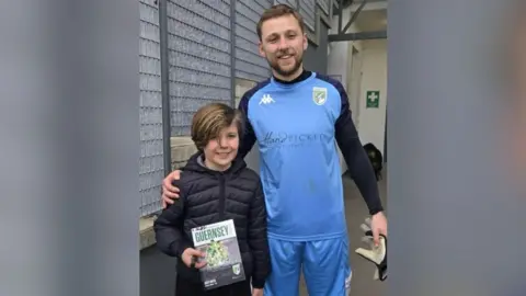 BBC Ciaran, wearing a black rain coat, is stood next to Josh Addison, who is wearing a light blue football kit with navy blue sleeves.