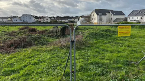 A green field with a fence in front of it which says on a yellow sign warning to public in red letters. 