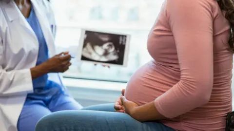A photo with the focus on the unrecognizable pregnant woman in the foreground as the unrecognizable doctor shows her an ultrasound on a digital tablet in the background