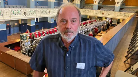 Alistair Ross from Oxford Brewery on the balcony at Oxford Town Hall. He is looking at the camera. Workers are setting up for the beer festival below.