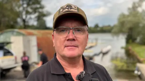 BBC Dr Derek Evans wears a black t-shirt and a grey baseball hat as well as a pair of rimless glasses. He is standing outside.