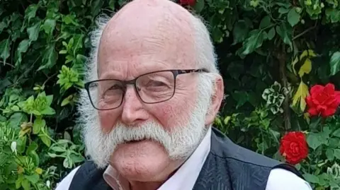 Doreen Savage Ray Savage smiles as he stands in front of a bush. He is largely bald with some grey hair on the side of his head and he has a grey beard. He wears glasses with a dark waistcoat and pink shirt.