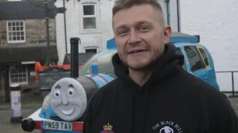 Alex Birkinshaw, a man with short hair and a stubble beard, standing in front of a - peaking from behind him - with the face of Thomas the Tank Engine constructed on its bonnet. The car has been sprayed blue with red stripes. Thomas's smiling face is stuck to the front with a funnel poking out the top and imitation wheels have been stuck to the sides of the car to complete the look.