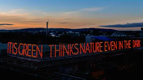 A photo of the neon sign reading "IT IS GREEN THINKS NATURE EVEN IN THE DARK" with a sunset in the background