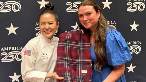 Kaci McEwan has long brown hair and is wearing a blue dress. Grace Pak has her dark hair tied up in a bun on top of her head. She is wearing a white outfit with the US Stars and Stripes flag on the sleeve. The women are holding a scarf, which has been woven in the US 250th anniversary tartan colours of red, blue and white.