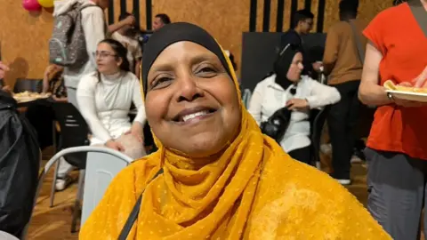 A woman wearing a yellow head scarf looking into the camera. Shew is smiling widely. There are many other people in the background of the picture. 