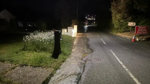Road at night with brown water flowing along it and a road closure sign to the right.