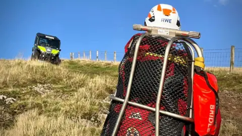 A person on a rescue stretcher is on a Scottish hillside with a vehicle waiting to take them to safety in the distance.