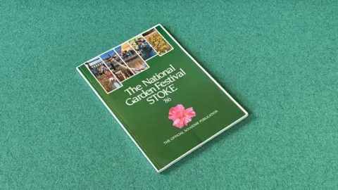 A brochure with a green background is positioned against a light green backdrop. The brochure contains images of flowerbeds and a large pink flower near the bottom.