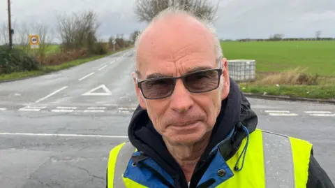 Roy Fabb is wearing glasses with tinted lenses and a hooded overcoat with a yellow hi-viz tabard on top. Behind him is a road junction with road signs.