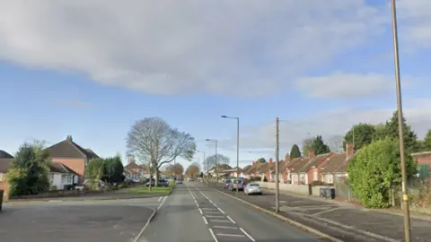 A long road with houses on either side and a large tree on the left. There are cars parked on the right 