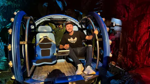 Sean Evans, wearing a cap, a black shirt, jeans and trainers, sitting on a theme park ride. 