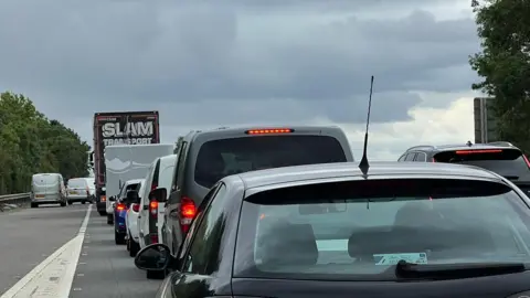 The dual carriageway of the A1 where a slip road is merging onto the carriageway. There are cars and lorries queuing on the road.