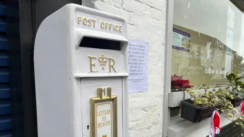 A white post box with the words "letters to heaven" written on it with decorated window and window boxes on the right