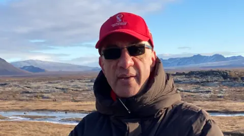 Keith Georgiou Keith Georgiou standing in front of a beach. They are wearing a red cap, sunglasses and a brown coat 