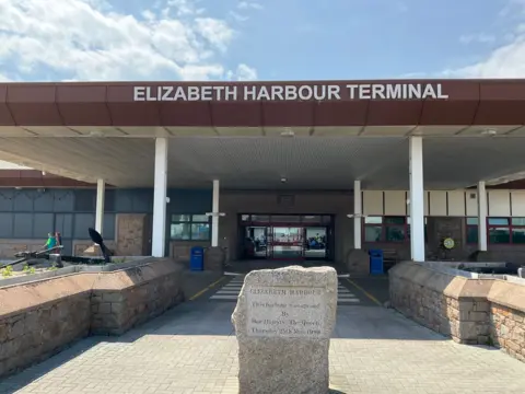 BBC Entrance to Elizabeth Harbour Terminal on a sunny day. An engraved rock says 'Elizabeth Harbour was opened by Her Majesty The Queen Thursday 25 May 1989'. Large glass double doors are open into the terminal.