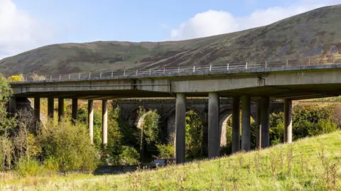 National Highways A view from the hillside at Tebay looking north-east towards two bridges carrying the M6 and the West Coast Mainline over the River Lune and in between the Howgill Fells just south of Tebay, Cumbria on a sunny day.