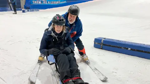 Margaret in a sled on the slopes of the Tamworth  Snowdome. A male staff member is on skis behind her is behind her