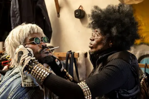TONY KARUMBA / AFP / GETTY IMAGES A man wearing studded clothing tends to a man wearing a blond wig and denim jacket.