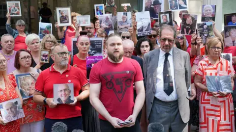 Matt Fowler speaks to the media while surrounded by other grieving family members holding up pictures of loved ones