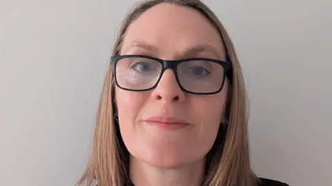 Head-and-shoulders portrait of a woman with shoulder-length, light brown hair wearing black glasses and a black top, standing against a plain light wall. She is wearing a name badge that reads Jayne Kerr, Practice Manager, with an iblu logo.