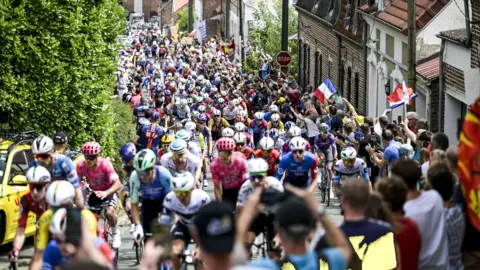 ASO & Charly López The Tour de France with a large crowd of cyclists climbing up a road. They are all wearing different colour jerseys and helmets. Crowds line the street, some holding up French flags. There are houses on the right side of the photograph.