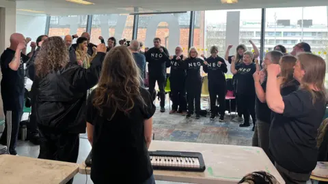 North East Opera choir rehearse their songs dressed in black t-shirts with the logo NEO. They are standing in a circle with their arms raised. In the foreground two women have their backs to camera next to a table with a keyboard on it.