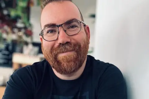 Daniel Allen A picture of Labour Councillor Daniel Allen. He is a man with short hair but he has a thick light brown beard and moustache and is wearing glasses. He is sat with a mug of coffee and his arms are folded. He has tattoos on his arms and is wearing a black t-shirt.