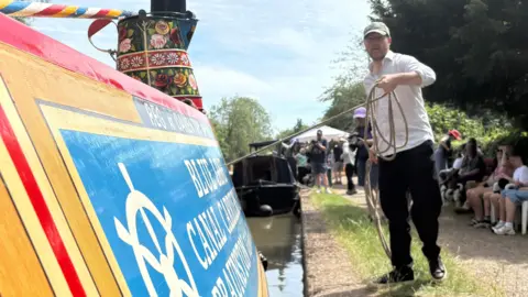 Martin Heath/BBC A brightly coloured canal narrowboat is moored alongside a path on a sunny day. The boat’s side, painted in red, yellow, and blue with decorative detailing, fills the left foreground. A tall, black chimney with gold bands and floral artwork rises from the boat, and part of a sign on the boat reads “B… Canal Cruis…” (partially obscured). On the right side of the image, Samuel West is standing on the towpath, holding a coiled rope that extends toward the boat. He is wearing a light-coloured shirt, dark trousers, and a cap, and is positioned slightly mid-step as if handling or pulling the rope.