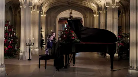 Kensington Palace A wide-shot of Catherine and Charlotte sitting and playing the piano. Charlotte is wearing a dark, long-sleeved top and tartan skirt. The hallway they are performing in has four visible Christmas trees, all decorated with red finishings and candles
