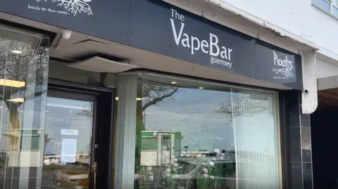 The picture shows the front of The VapeBar in Guernsey. The shop sits on the ground floor of a white building, and the black sign above the entrance reads “The VapeBar guernsey” in white lettering. 
