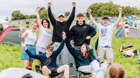 Izzy Challoner / Truck Festival Campers in the campsite cheer, with their arms aloft. Two at the front are sat on camp chairs, the five behind are standing.