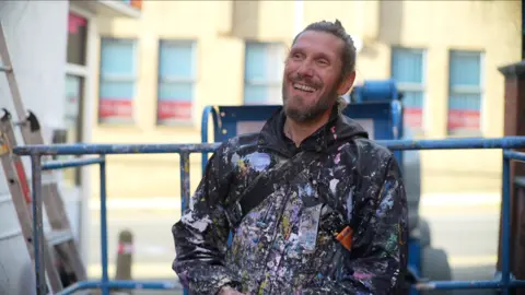 Jamie Niblock/BBC Artist Atma laughs as he looks away from the camera. He stands in front of a cherry picker that rests near the bottom of the building. He wears a black coat that is splattered with different coloured paint. He has long brown hair that has been tied up in a bun on top of his head and he has a thick dark beard.