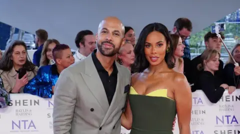 PA Media Marvin Humes with short dark beard, looking at the camera and wearing a grey jacket over a black shirt. He is standing next to Rochelle who has medium-length straight dark hair, wearing a green dress with yellow tips. There are several people behind a NTA banner behind.