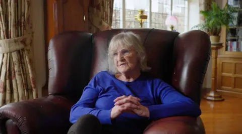 Mary Lanigan, the former leader of Redcar and Cleveland Council, sits in a large, reddish-brown leather armchair. She wears a blue long sleeve top and glasses. She has shoulder length grey hair and sits with her legs crossed and her fingers linked together on her lap.