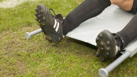 Getty Images A person's legs lying on a grey stretcher on the grass. The person is wearing black knee-socks and black football boots with two white stripes on the side of each boot. 